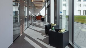 Un couloir d’hôtel moderne, doté de luminaires circulaires au plafond, de murs en verre et d’un espace détente offrant une vue sur la façade de l’immeuble.