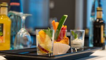 A tray with a fruit cocktail, a cupcake, and a small glass of a creamy beverage, set against a backdrop of various bottles and glasses.