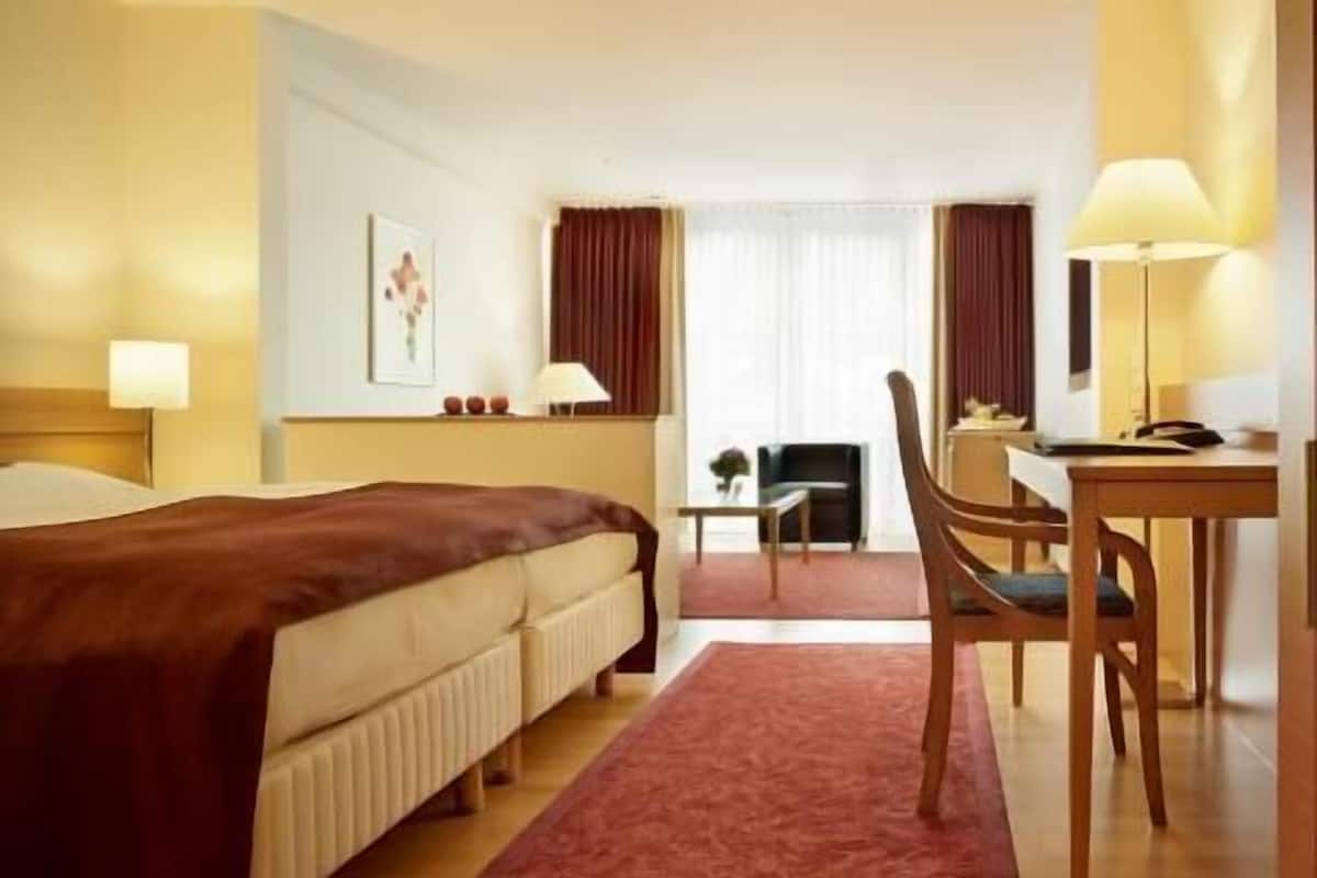 Ein Hotelzimmer mit einem Bett, einem Schreibtisch, einem Stuhl, einer Lampe und einem fenster mit Vorhängen.