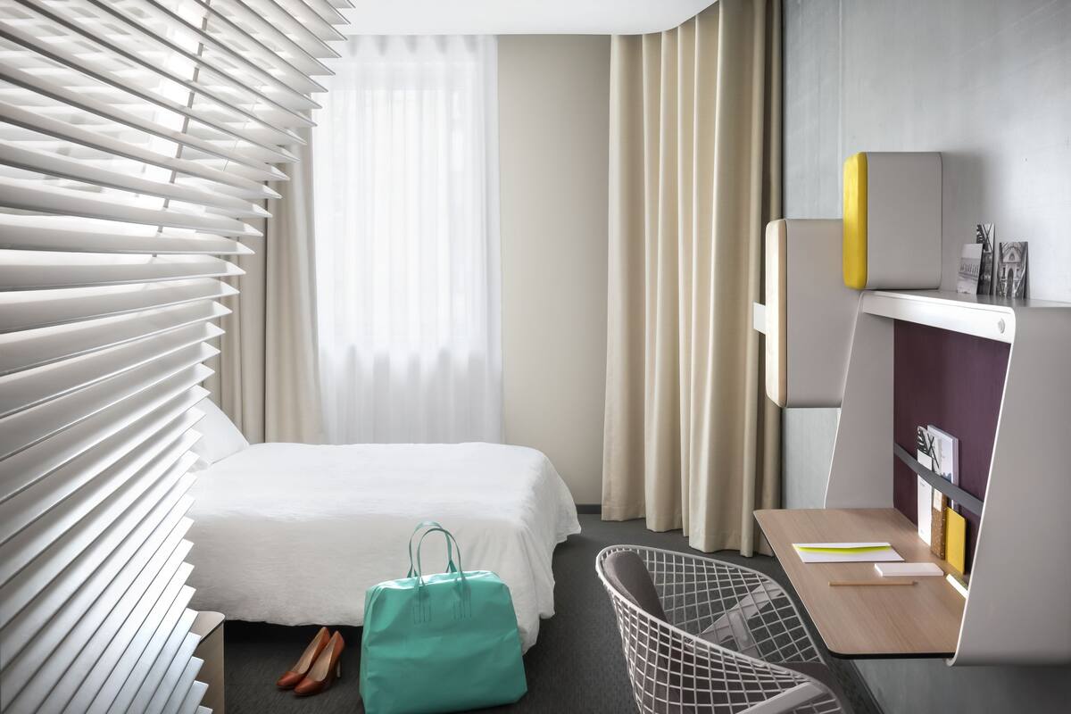 Una habitación de hotel con cama, un escritorio y silla, una ventana con persianas, y una mesita con una bolsa y un cuaderno.