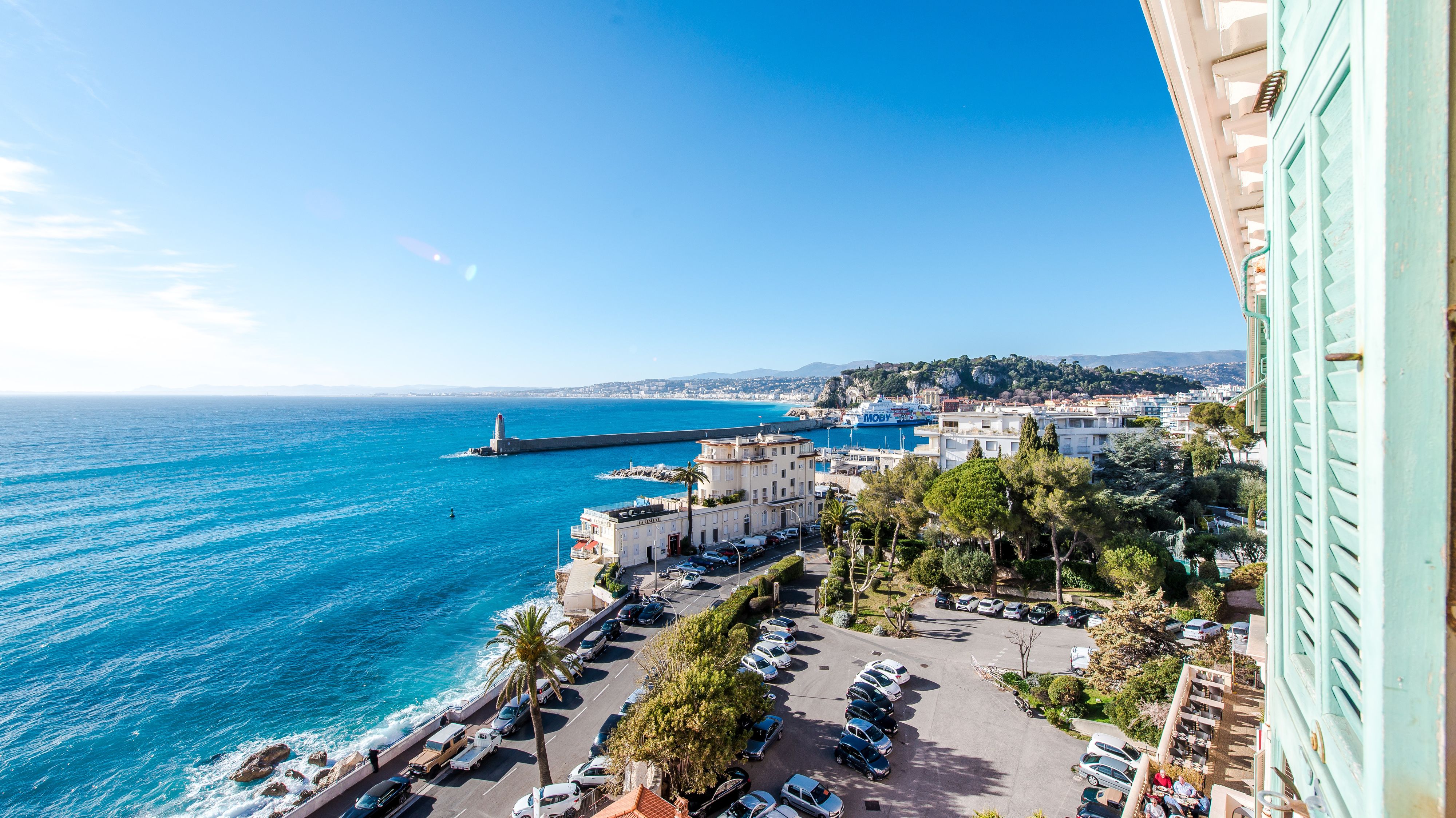 A coastal cityscape with a view of the sea, a lighthouse, and a parking area with cars.