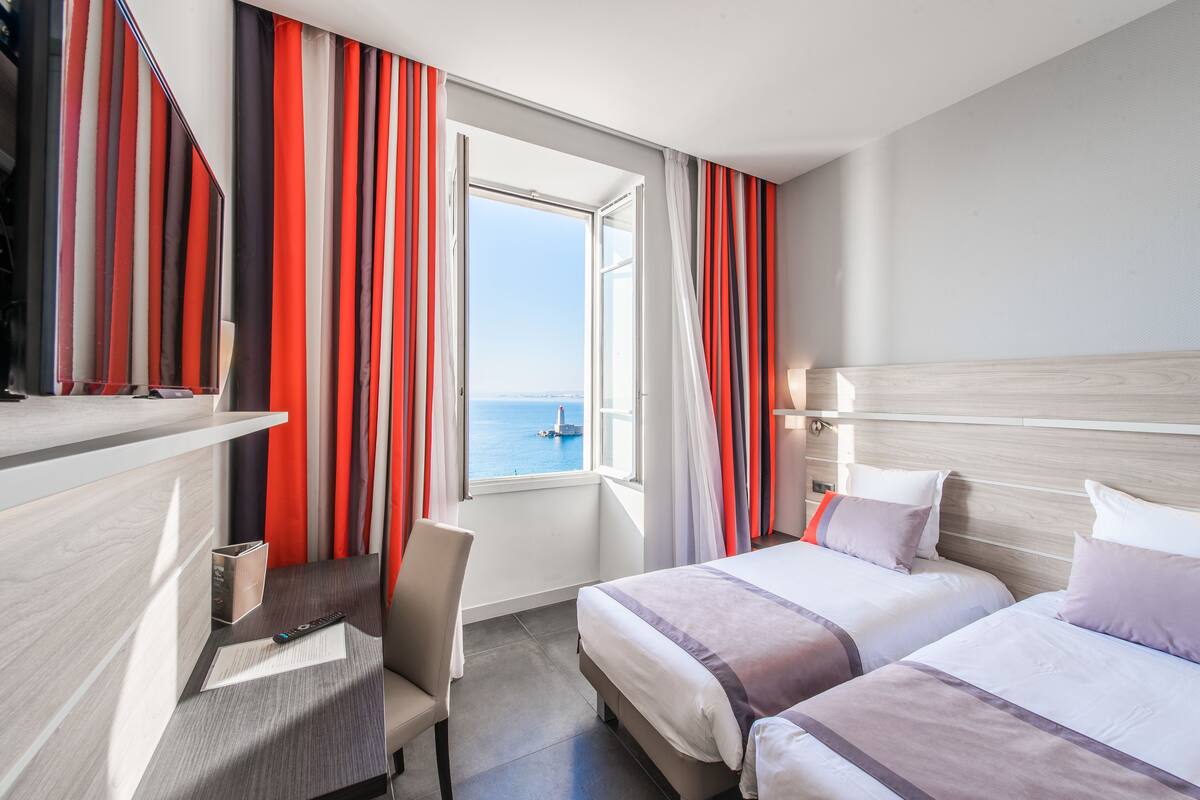 A hotel room with two beds, a desk, and a view of the sea.