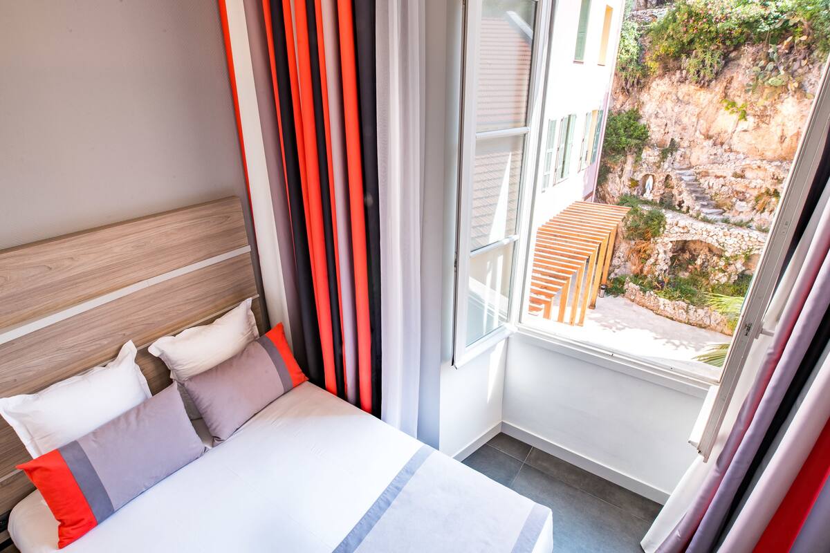 A hotel room with a bed, a window with curtains, and a view of a rocky outdoor area.