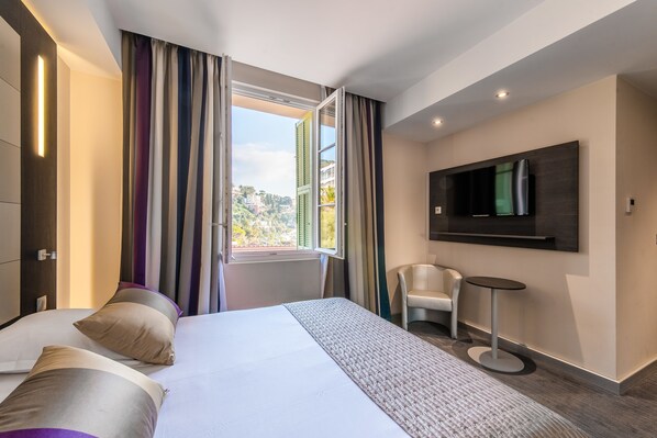 Une chambre d’hôtel moderne avec un grand lit, un téléviseur fixé au mur, une petite table ronde et une vue sur la ville par la fenêtre.