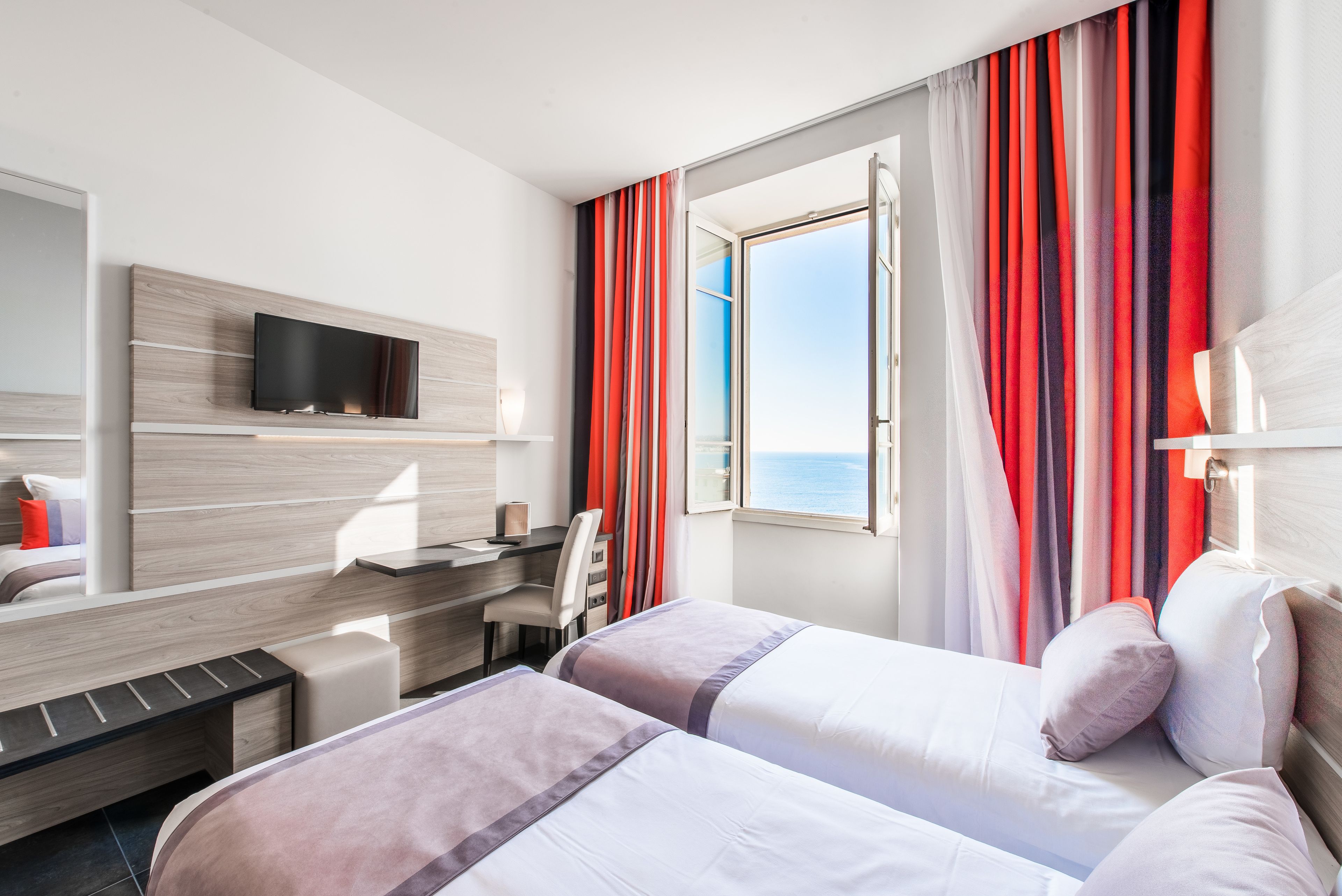 A hotel room with two beds, a desk, a TV, and a window with curtains.