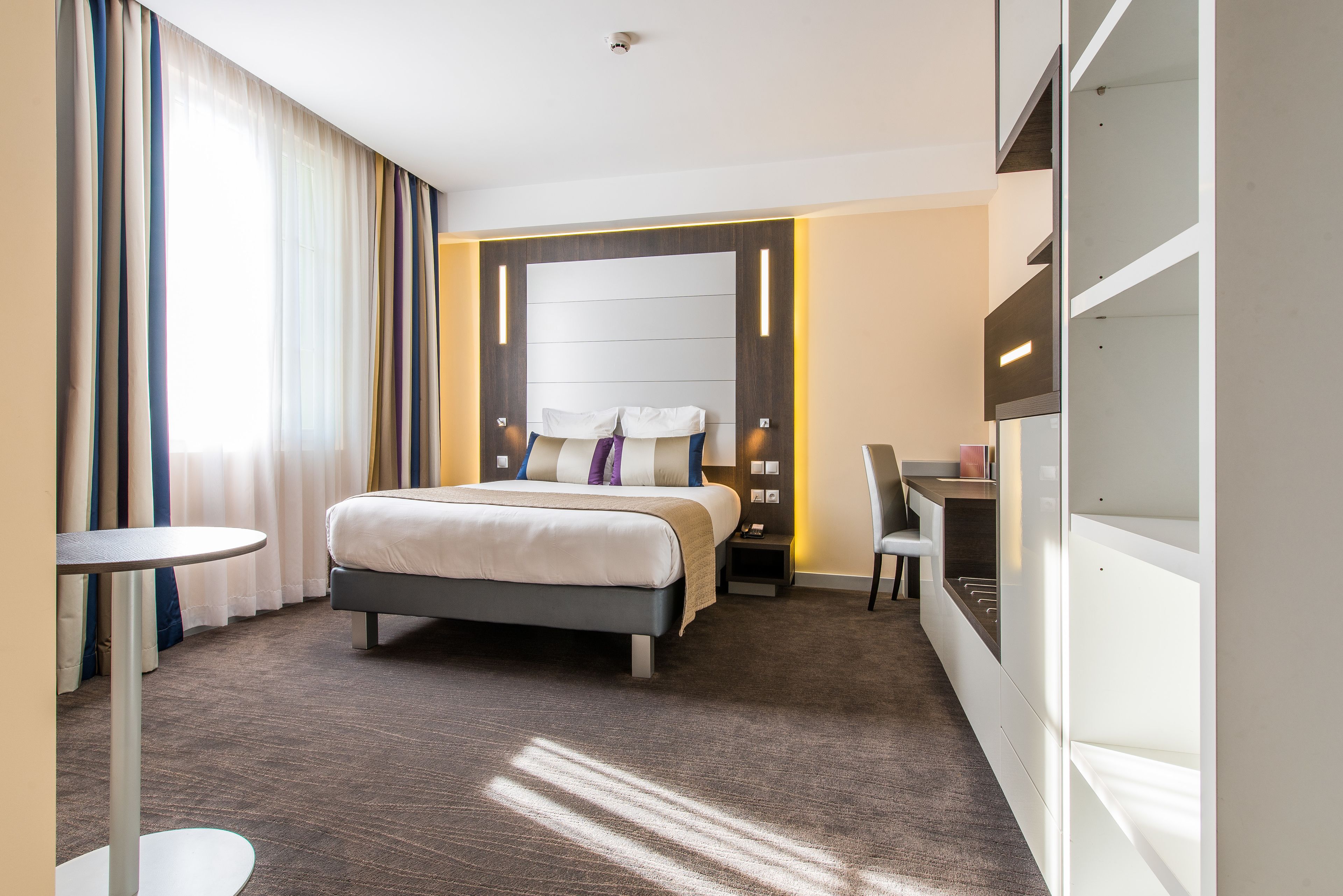 A hotel room with a bed, bedside table, chair, desk, and built-in wardrobe.