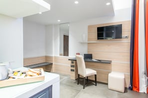 A modern hotel room with a kitchenette, a flat-screen TV, a comfortable chair, and a small table with breakfast items.