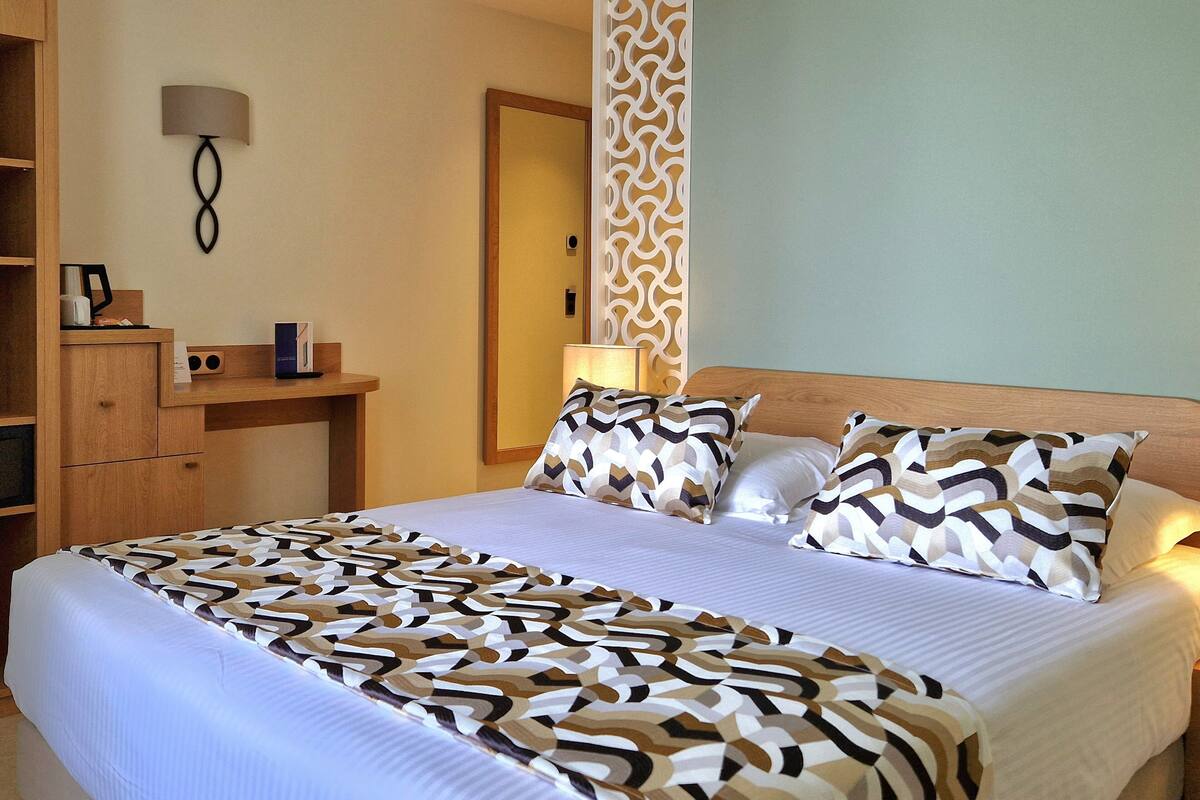 A hotel room with a large bed, a wooden headboard, a bedside table with a lamp, and a decorative wall panel.