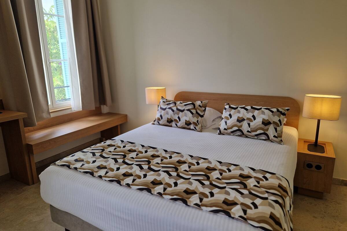 A neatly made bed with patterned pillows, a bedside table, a nightstand with a lamp, and a window with curtains.