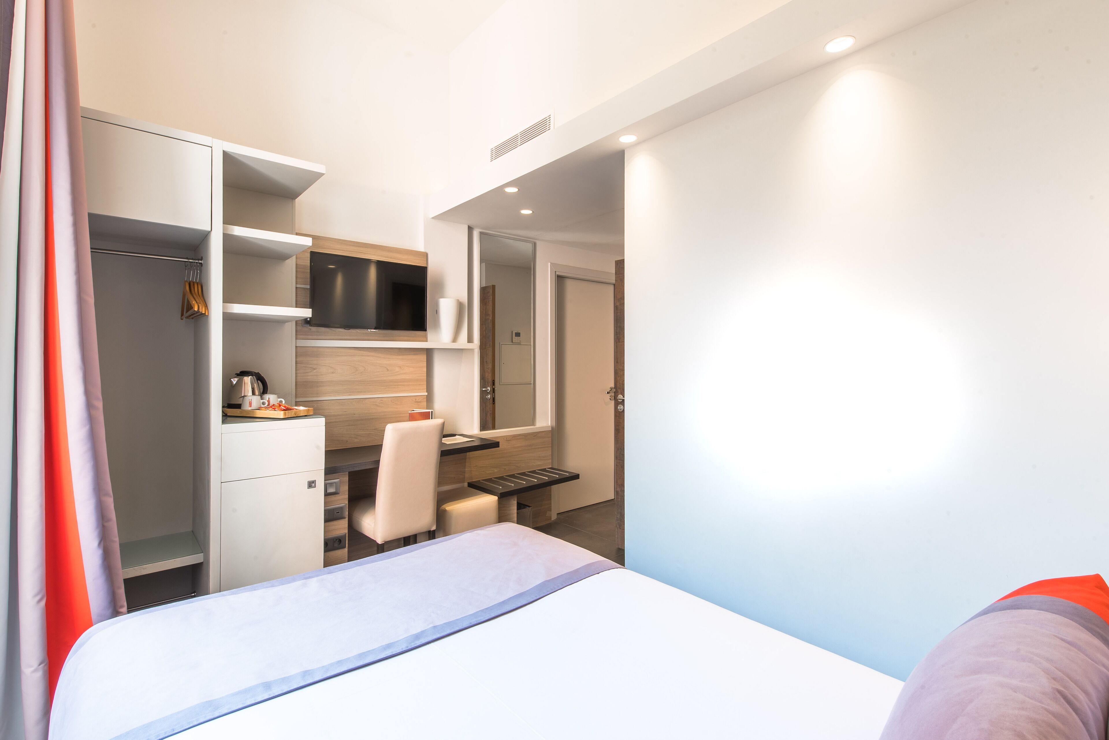 A modern hotel room with a bed, a desk, a chair, and a wardrobe.