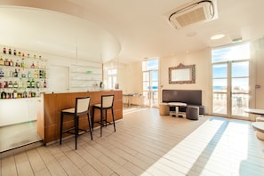 A modern bar area with a wooden counter, two bar stools, and a variety of liquor bottles. There is a seating area with a sofa and a table, and large windows offering a view of the outdoors.