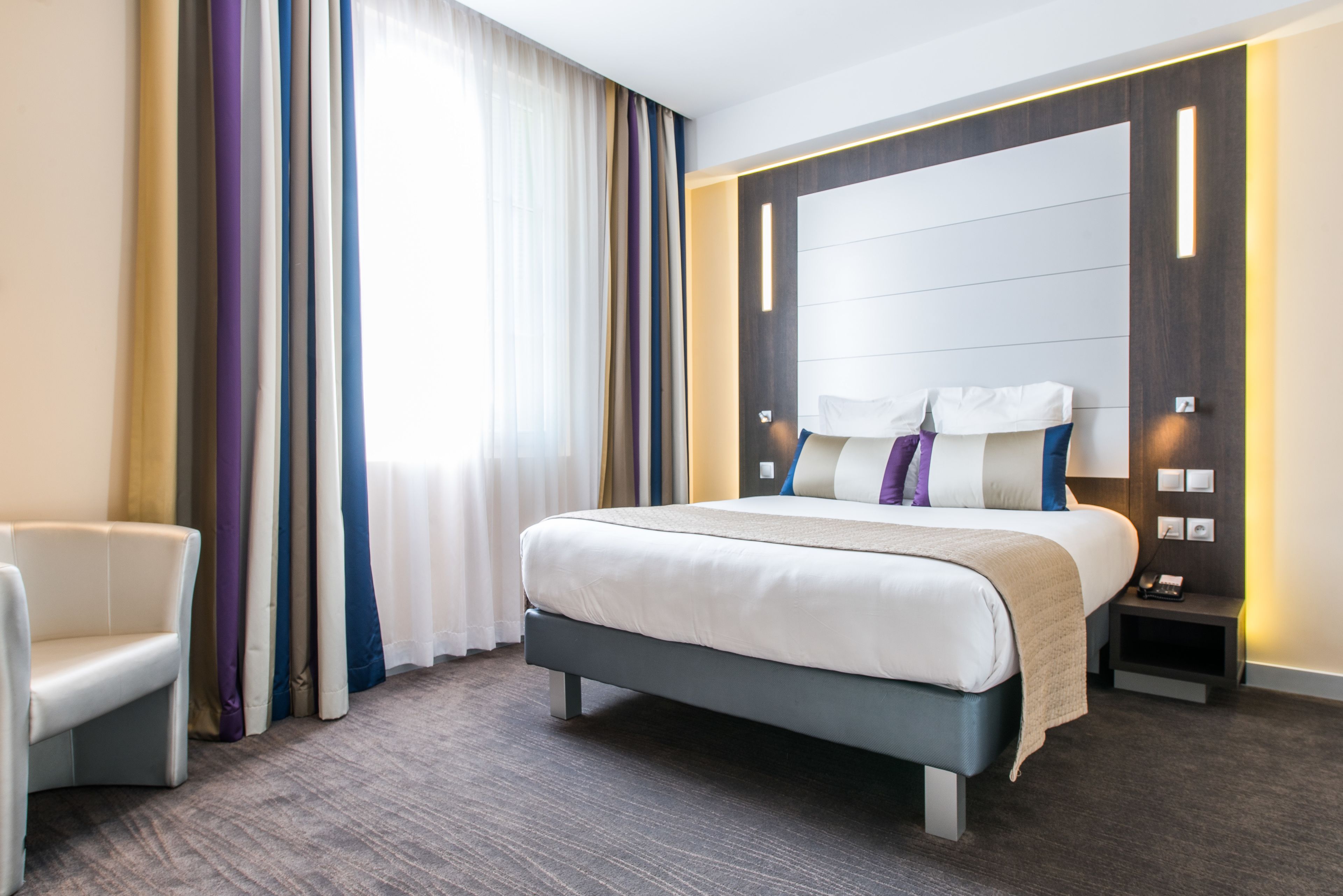 A modern hotel room with a large bed, a bedside table, a chair, and a view of the city through the window.