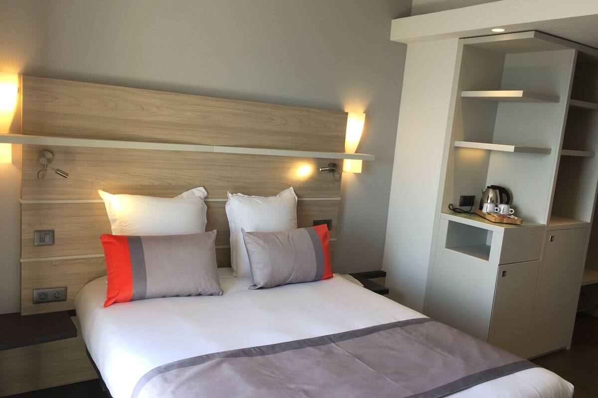 A hotel room with a large bed, a wooden headboard, bedside lamps, a small desk with a kettle, and a built-in wardrobe.