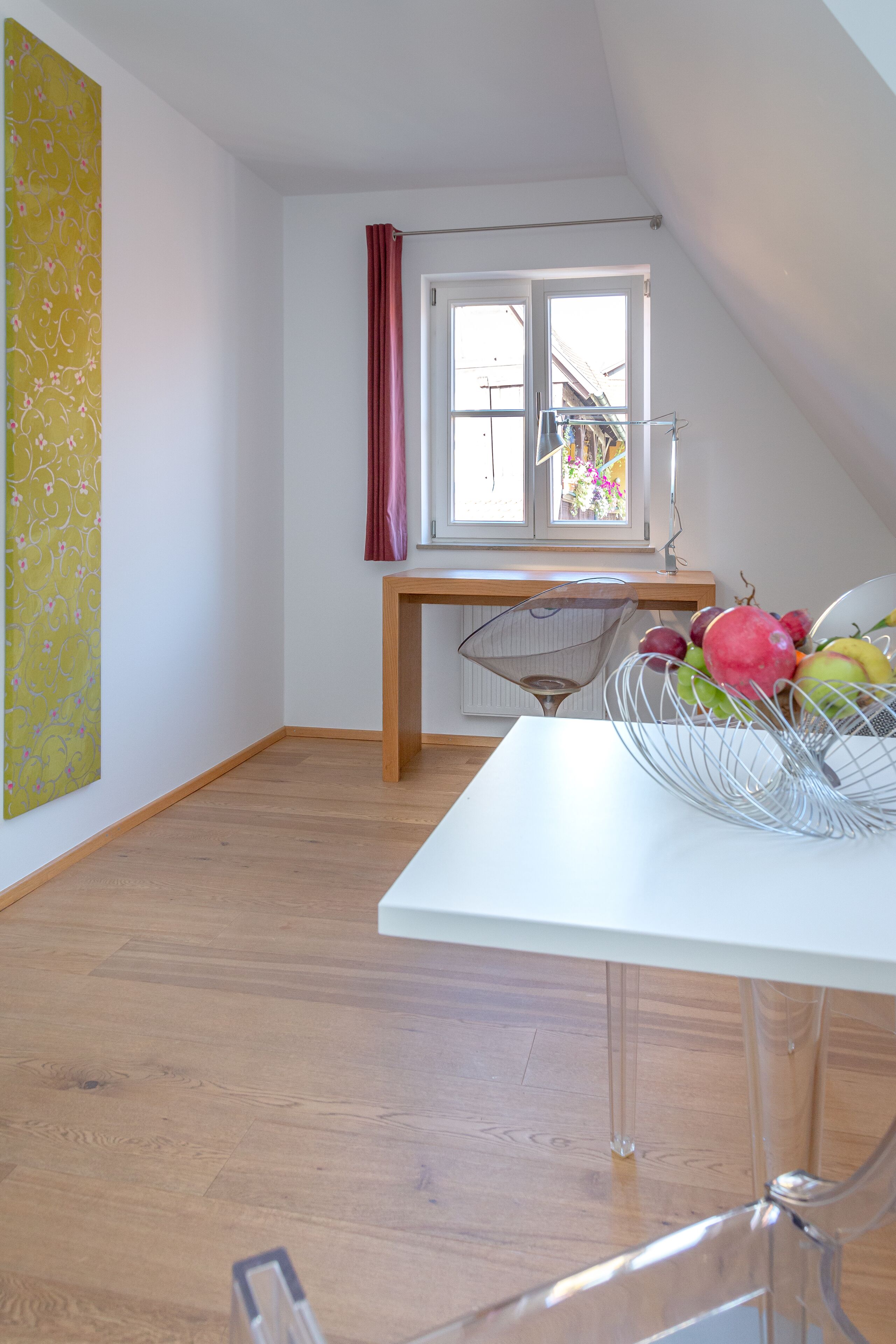 Een kamer met een witte tafel, een metalen draadmanden gevuld met fruit, een houten bureau en een raam met een rode gordijn.