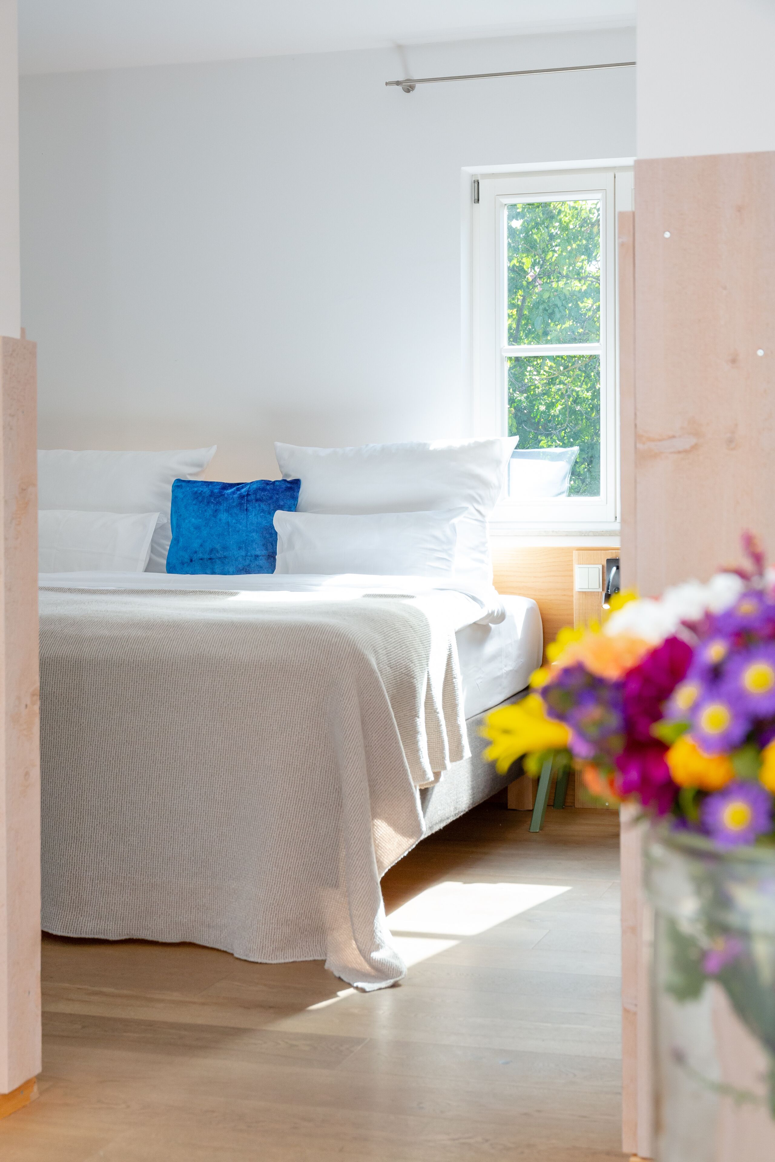 A bedroom with a bed, white bedding, a blue pillow, a window with a view of trees, and a vase with flowers.