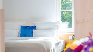 A bedroom with a bed, white bedding, a blue pillow, a window with a view of trees, and a vase with flowers.