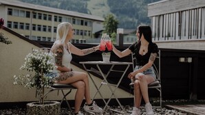 Dos personas sentadas en una mesa con bebidas, al aire libre con edificios y montañas al fondo.
