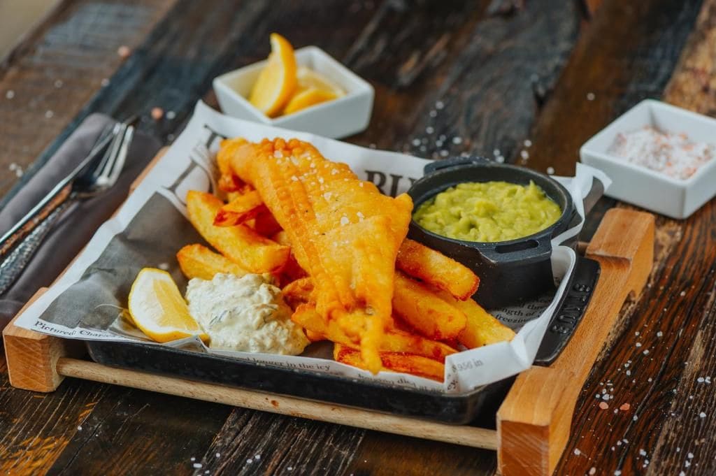 Uma bandeja com peixe frito, batatas fritas, fatias de limão, molho tártaro e ervilhas amassadas.