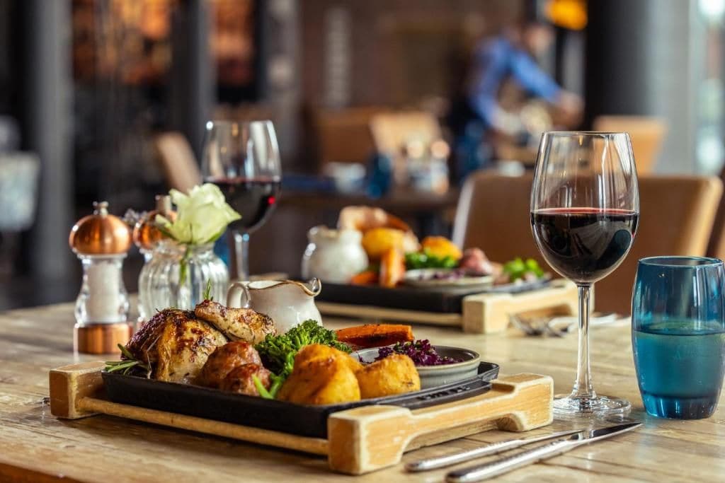 A dining table with a roasted chicken, vegetables, and a glass of red wine.