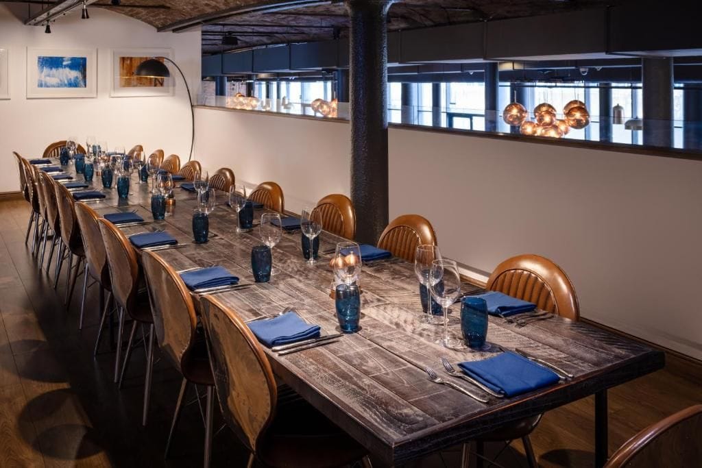 Una larga mesa de comedor dispuesta para una comida, con sillas, suelo de madera y un ventanal con vistas.