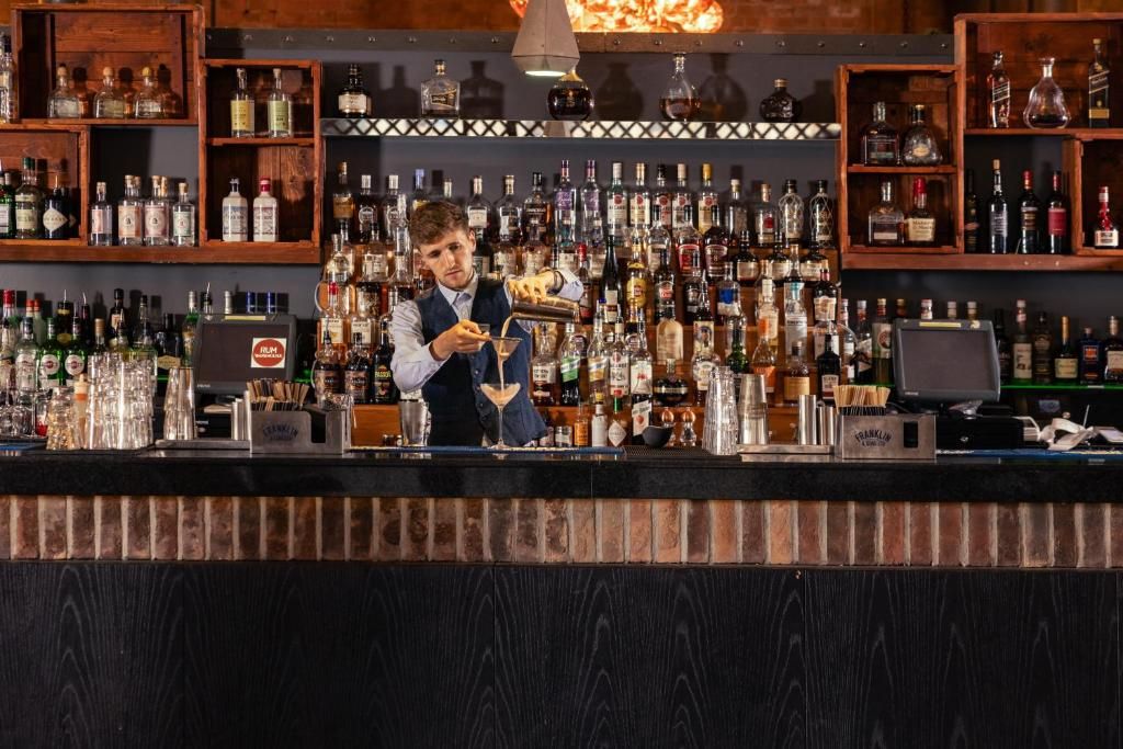 A bar with a bartender pouring a drink, shelves of liquor bottles, and a cash register.
