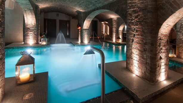 An indoor swimming pool with stone arches and a central fountain, surrounded by stone walls and illuminated by lanterns. - Titanic Hotel Liverpool (Liverpool)
