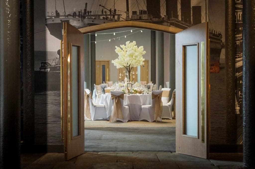 Un comedor interior con mesas dispuestas para una comida, un gran centro de mesa floral y vistas a un barco a través de un arco.