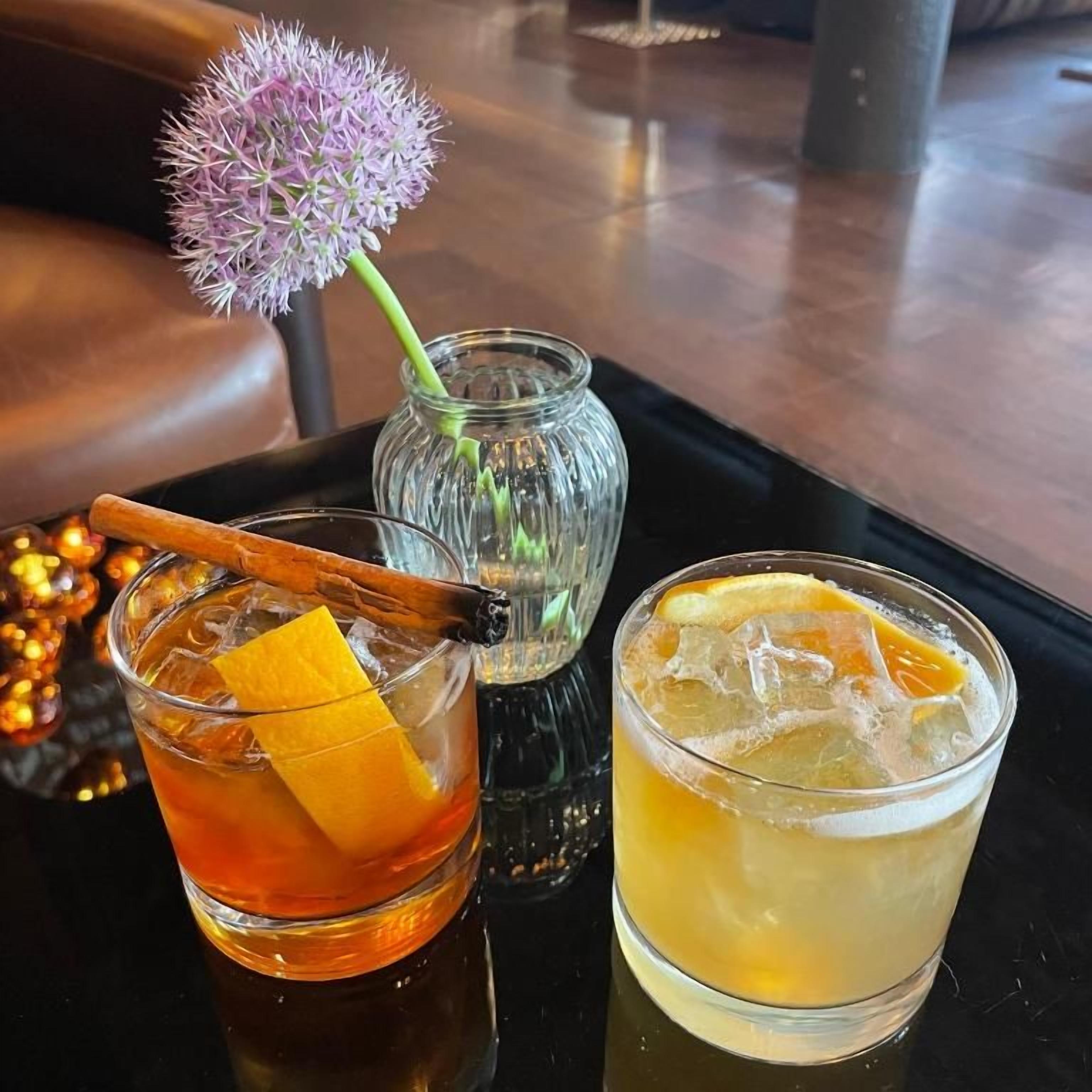 Two glasses of beverages on a tray, one with a cinnamon stick and orange peel, and the other with ice and a slice of orange.