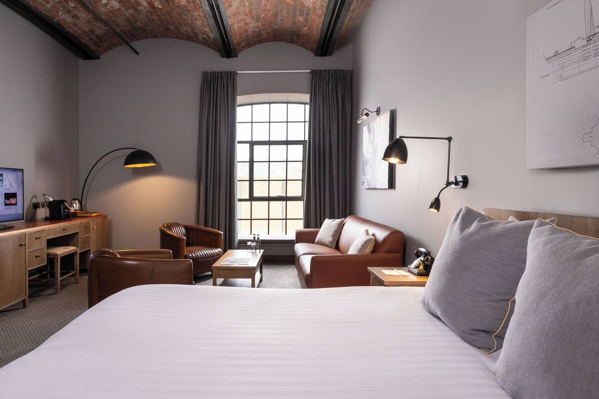 A hotel room with a large bed, a desk with a computer, a chair, a sofa, and a window with curtains.