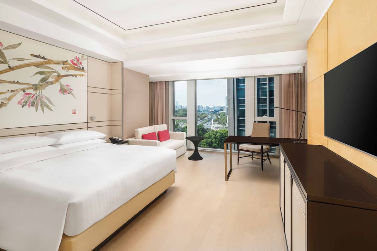 A hotel room with a large bed, a desk, a chair, a TV, and a view of the city through large windows.