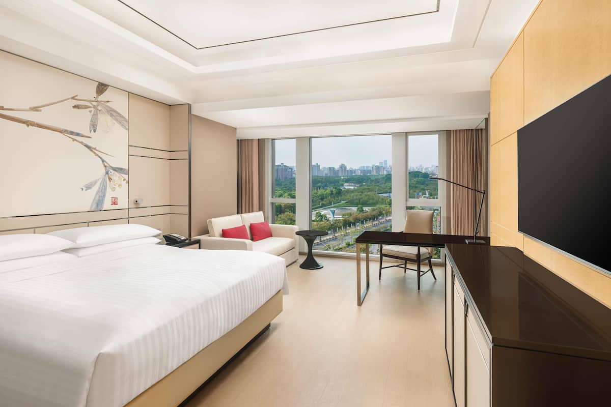 A modern hotel room with a large bed, a desk, a chair, a TV, and a city view through large windows.