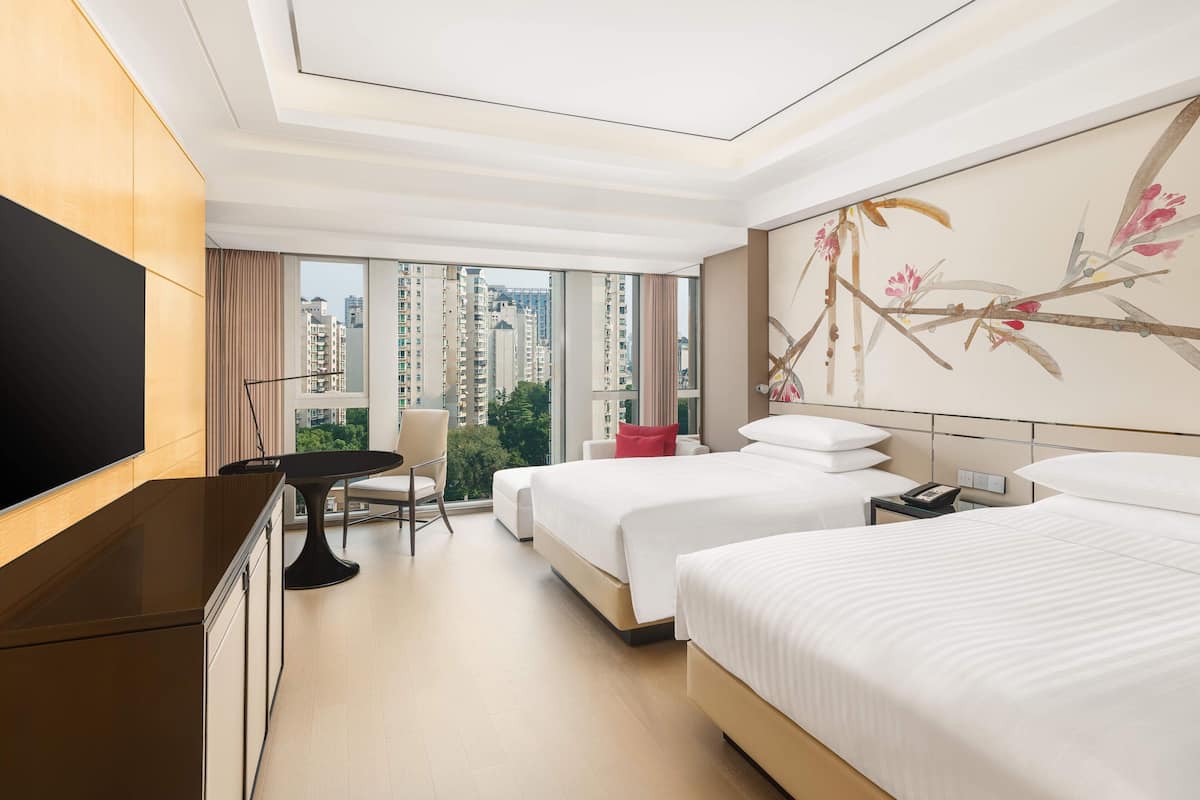 A hotel room with two beds, a TV, a desk, and a large window with a city view.