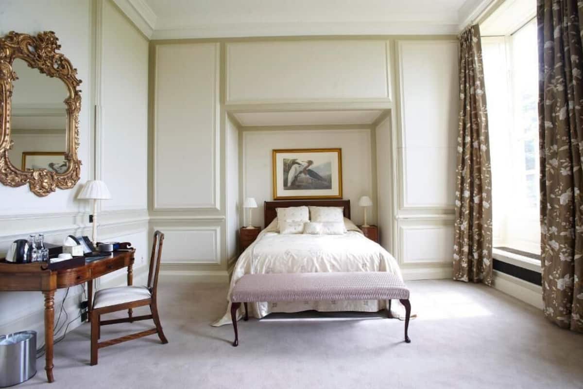 A bedroom with a large bed, a desk with a chair, a mirror, and a window with curtains.