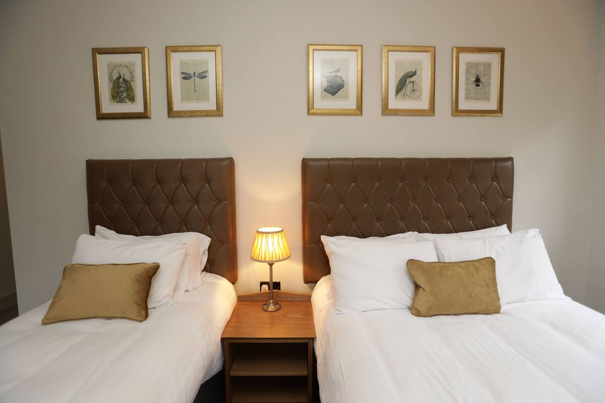A double room with two beds, a nightstand with a lamp, and framed artwork on the wall.