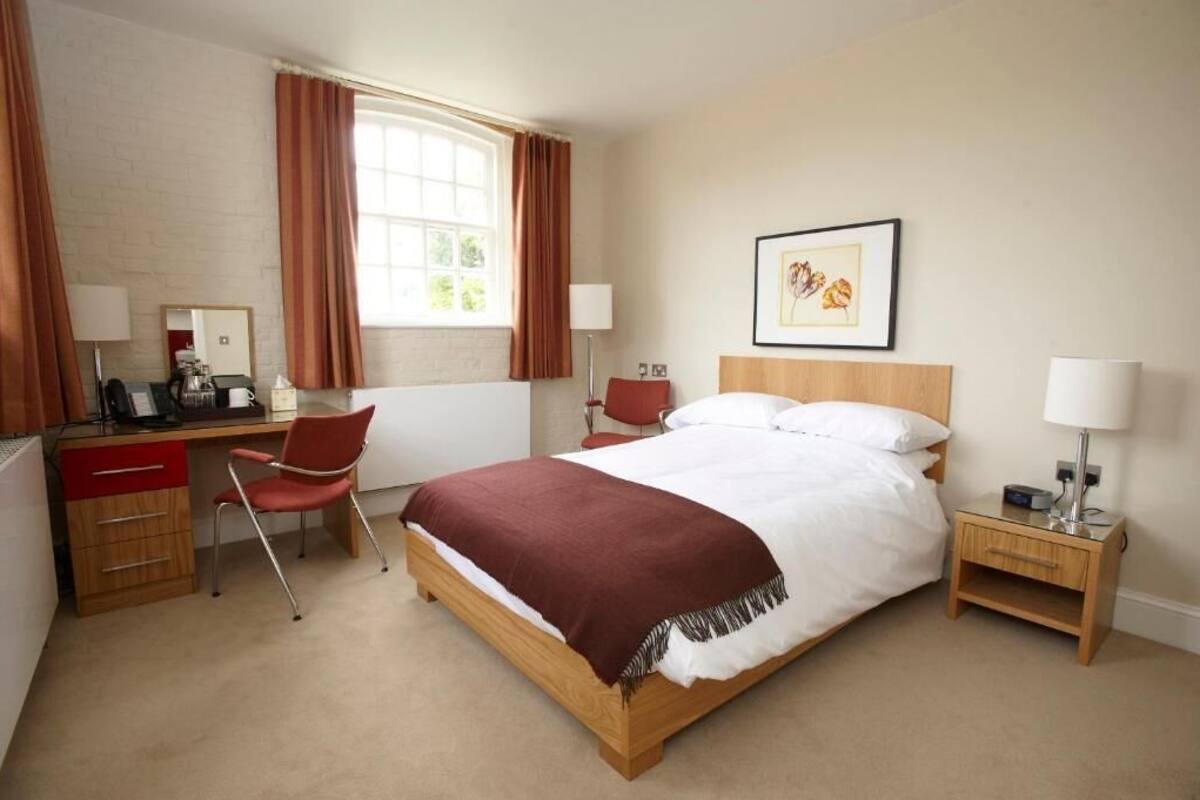 A hotel room with a wooden bed, a desk with a chair, a nightstand, and a window with curtains.