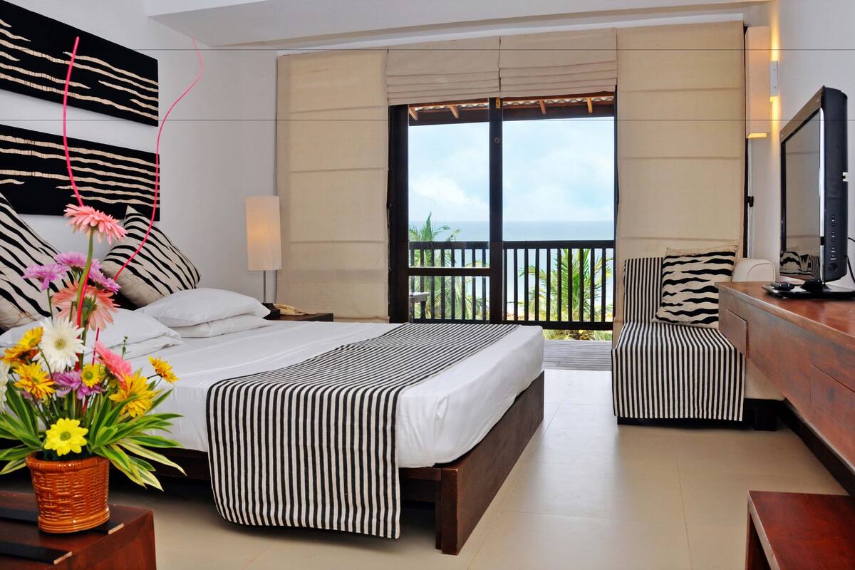 A hotel room with a bed, a chair, a TV, and a view of the ocean.