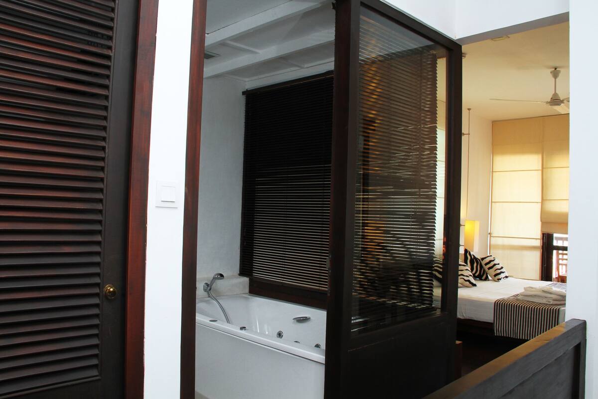 A room with a bathtub, a bed with striped bedding, and a window with blinds.