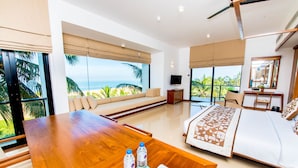 A modern hotel room with a large wooden table, a bed with patterned bedding, a sofa, and a TV. - Goldi Sands Hotel (Negombo)