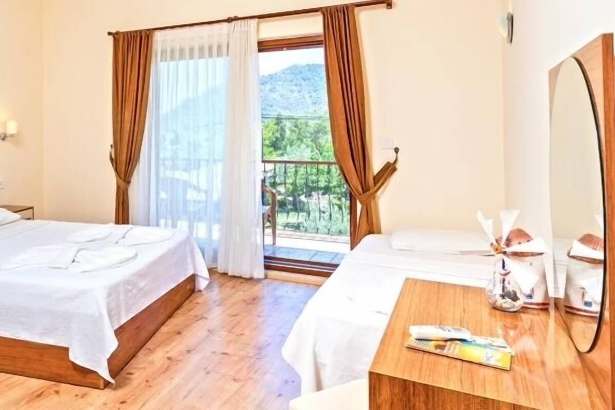 A hotel room with two beds, a wooden floor, a balcony with curtains, a small table with toiletries, and a large mirror.