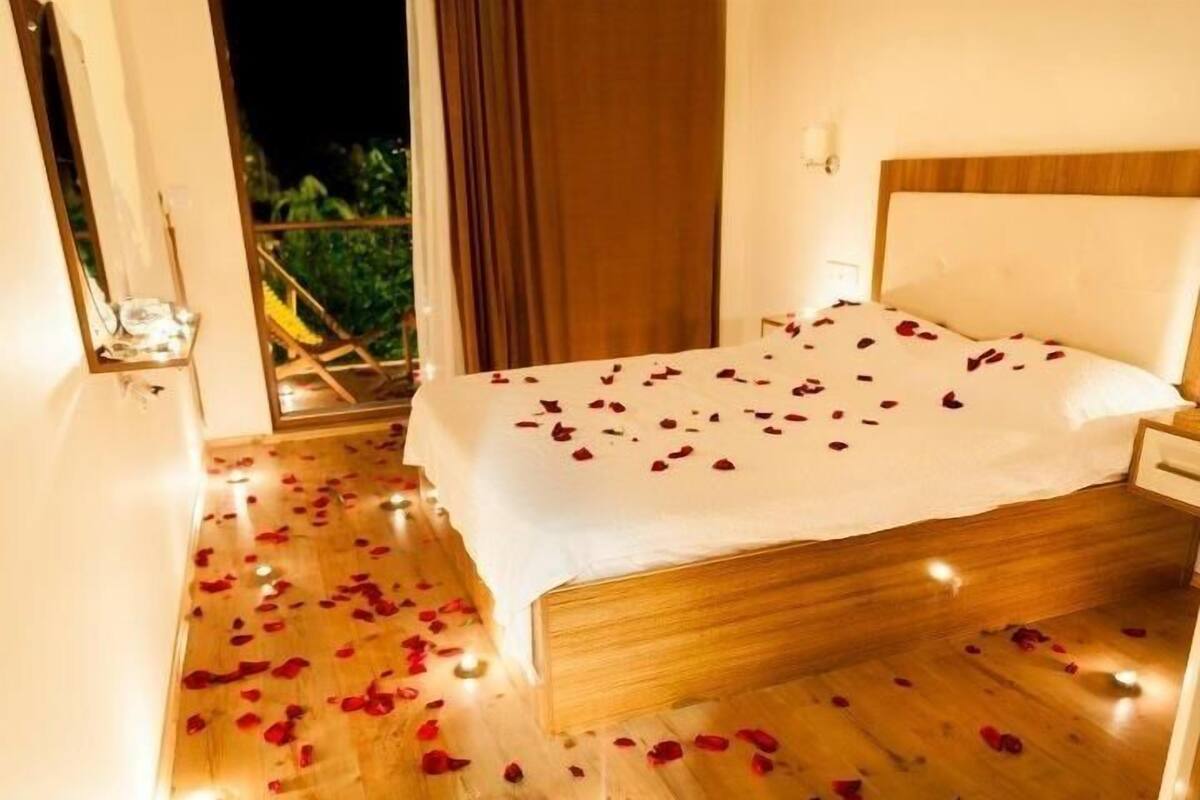 A hotel room with a bed covered in rose petals, a wooden floor, and a view of greenery outside.
