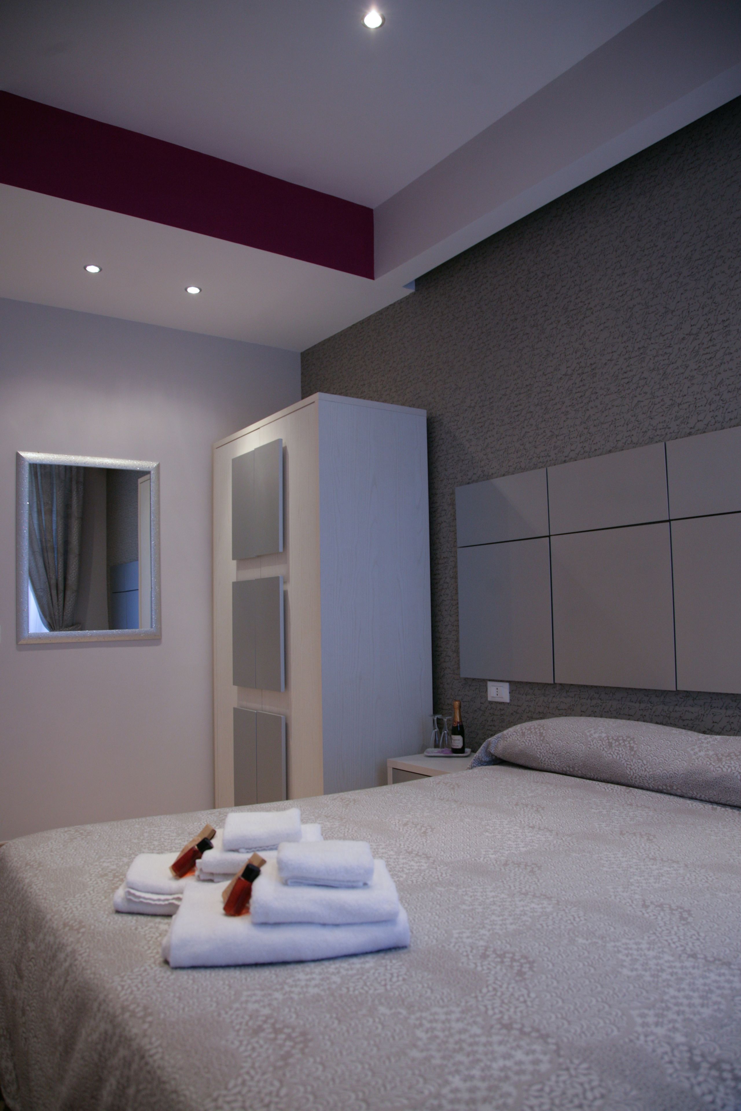 A modern hotel room with a neatly made bed, a wardrobe, a bedside table with a bottle, and a large window with curtains.