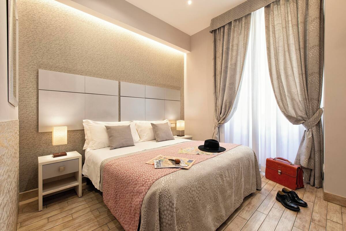 Un dormitorio ordenado con una cama grande, lámparas de noche, una mesita de noche, una maleta roja y un par de zapatos en el suelo.