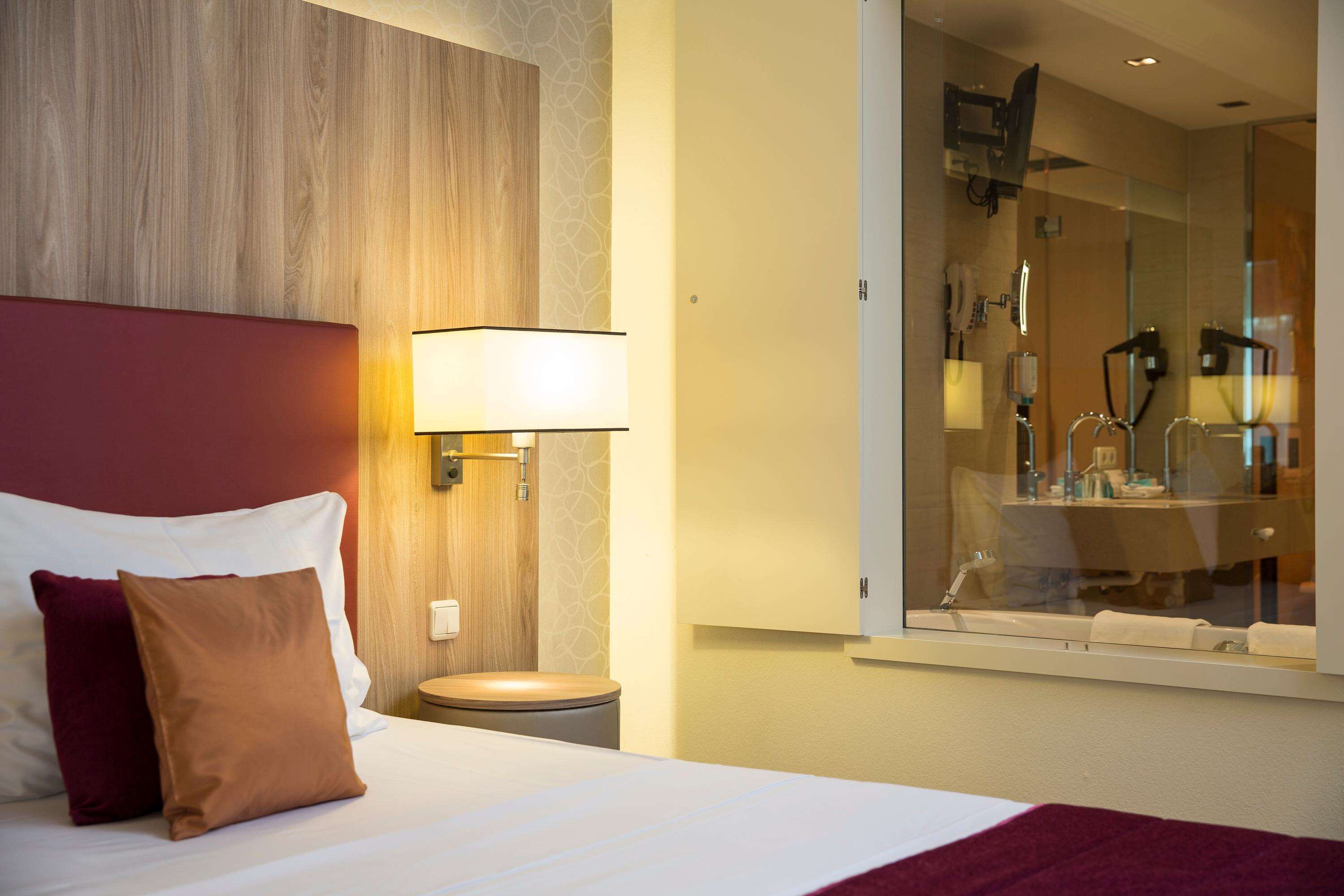 Habitación de hotel con cama, mesita de noche, lámpara y baño visible a través de un panel de vidrio.