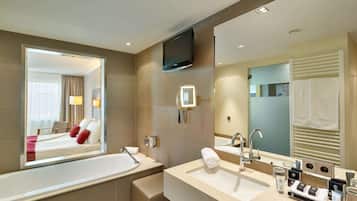 A modern bathroom with a large mirror, a bathtub, a sink, and a flat-screen TV mounted on the wall.