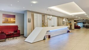 A modern hotel reception area with a white reception desk, red seating, and a large window with curtains. - Hotel Schillerpark Linz, a member of Radisson Individuals (Linz)