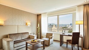 A hotel room with a sofa, two armchairs, a wooden coffee table, and a desk with a lamp. There is a large window offering a city view. - Hotel Schillerpark Linz, a member of Radisson Individuals (Linz)