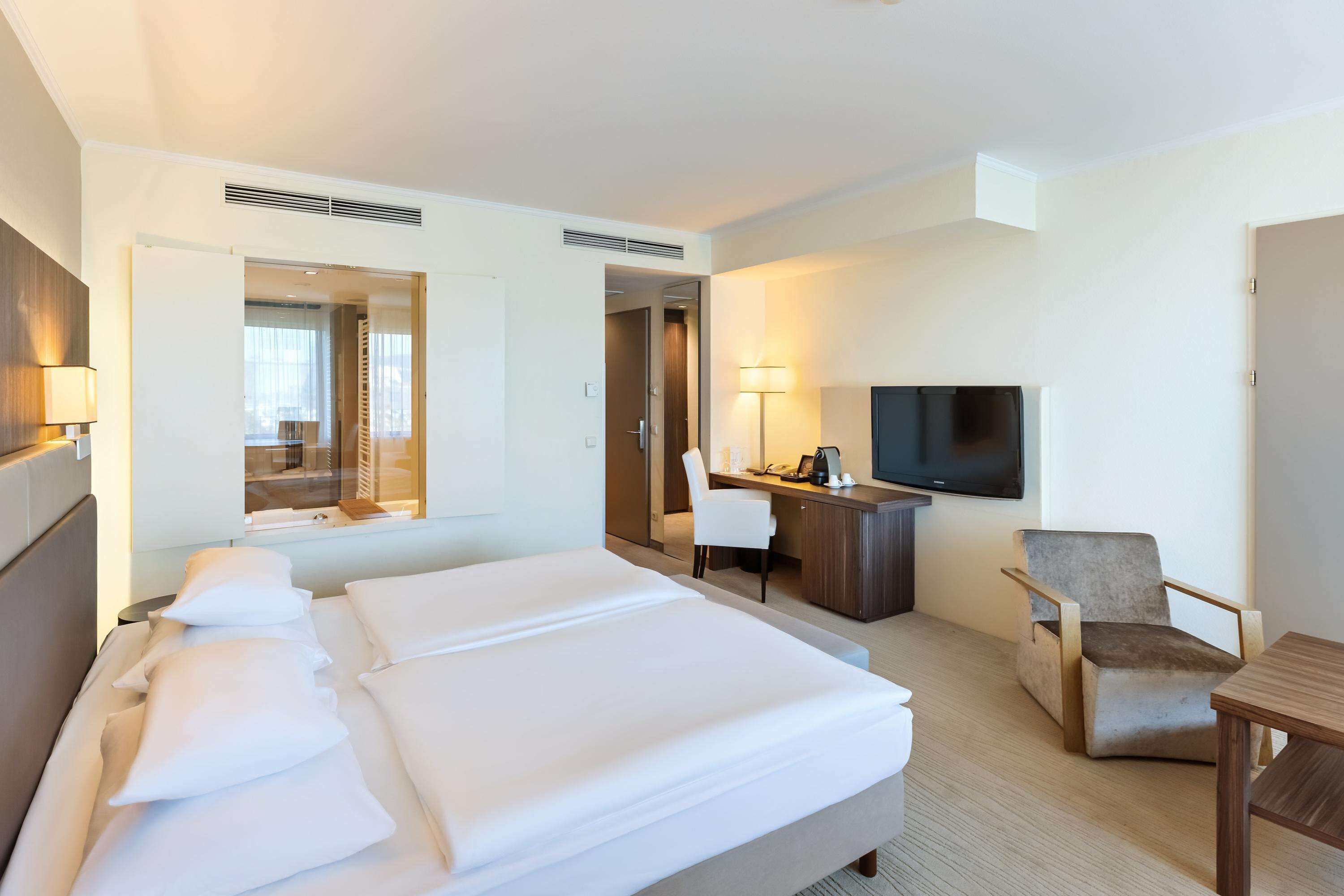 A hotel room with a large bed, a desk, a chair, a TV, and a window.