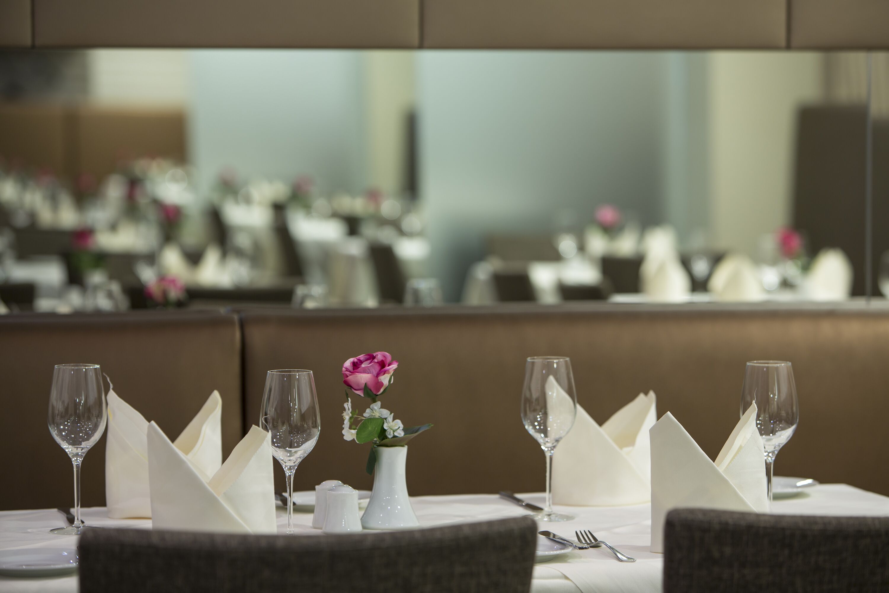 A restaurant setting with a table set for dining, including wine glasses, a vase with a flower, and neatly folded napkins.