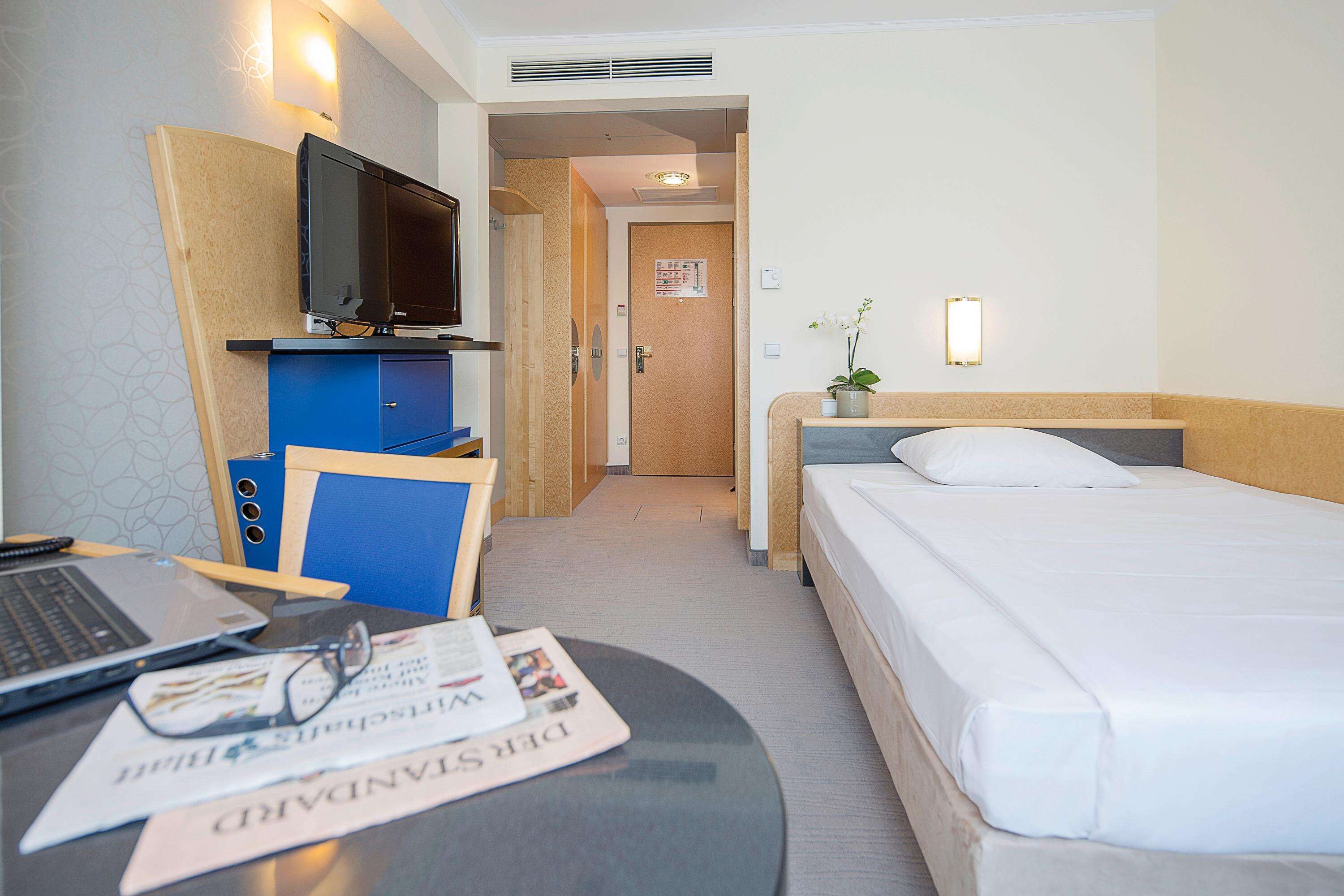 Habitación de hotel con cama, escritorio, laptop, televisión y una pequeña planta en maceta.
