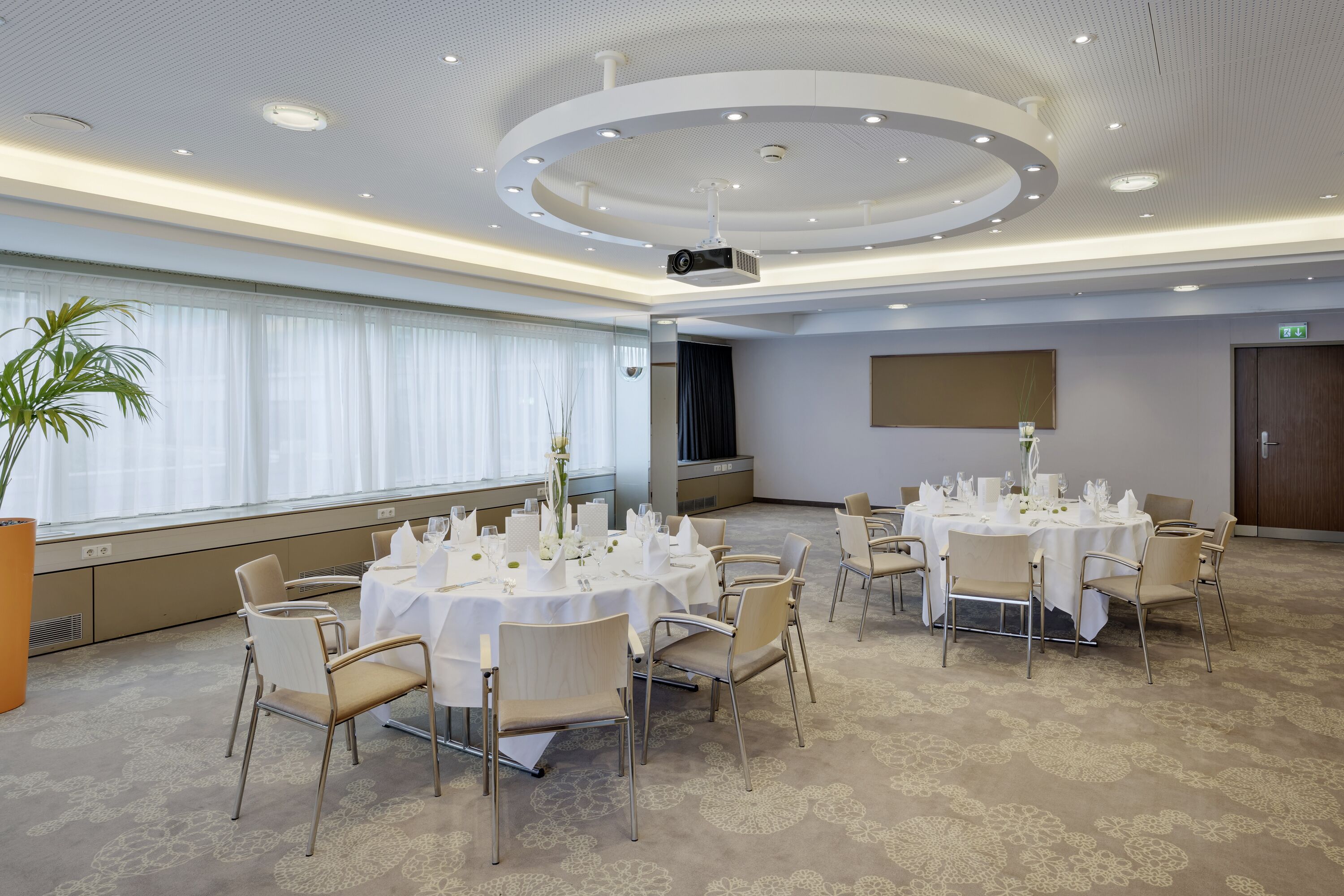 A conference room with round tables, chairs, a projector, and a large window with curtains.